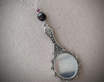 May include: Silver-toned pendant necklace featuring a miniature hand mirror design. The mirror has an ornate handle and a decorative frame. A black faceted bead and a small purple bead are suspended above the mirror. The necklace chain is delicate.
