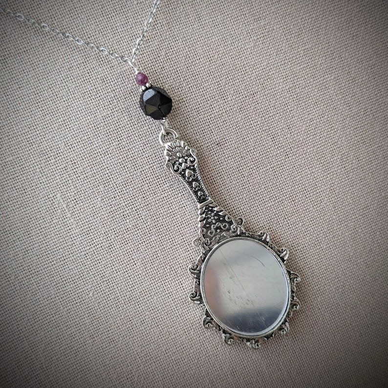 May include: Silver-toned pendant necklace featuring a miniature hand mirror design. The mirror has an ornate handle and a decorative frame. A black faceted bead and a small purple bead are suspended above the mirror. The necklace chain is delicate.