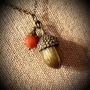 May include: A gold acorn pendant necklace with a red gemstone accent. The necklace is on a brown fabric background.