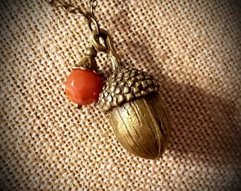 May include: A gold acorn pendant necklace with a red gemstone accent. The necklace is on a brown fabric background.