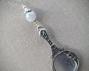 May include: A silver-toned pendant necklace featuring a small, hand mirror design. The pendant has intricate detailing and a round mirror. A white, speckled bead hangs above the mirror, connected by a delicate chain.