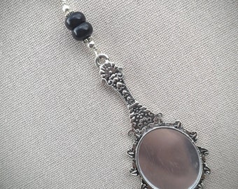 May include: A silver-toned pendant necklace featuring a miniature hand mirror design. The mirror has an oval shape with an ornate, decorative frame. Two black beads are strung on the chain above the mirror.