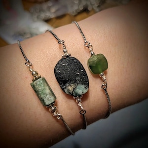 May include: Three bracelets with silver chains and green and black stone beads. The bracelets are layered on a person's wrist.