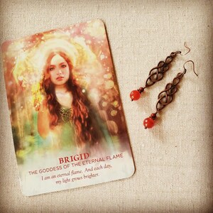 May include: A card with a painting of a woman with red hair and a green dress. The card has the text "Brigid The Goddess of the Eternal Flame I am an eternal flame. And each day, my light grows brighter." A pair of copper earrings with red stones hang on a beige fabric background.