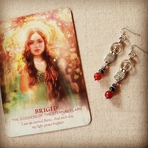 May include: A pair of silver earrings with a spiral design and red, green, and purple beads, next to a card with the text "BRIGID THE GODDESS OF THE ETERNAL FLAME". The card features an illustration of a woman with long brown hair.