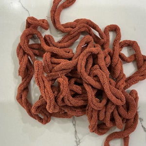 May include: A pile of twisted, rust-colored fabric cords. The cords are thick and appear to be made of a knitted or woven material. The texture is visible, and the cords are arranged in a tangled, organic pattern against a white marble surface.