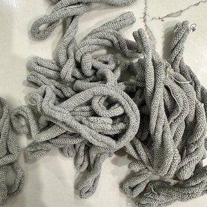 May include: A collection of gray knitted cords, tightly twisted and looped, creating a tangled arrangement. The cords have a textured appearance, with a mix of light and dark gray threads. The background is a light-colored surface.