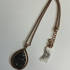 May include: A copper-toned necklace with a teardrop pendant filled with dark gray crystals. The pendant is framed with a gold-colored border and hangs from a matching chain. A tag with text is attached to the chain.