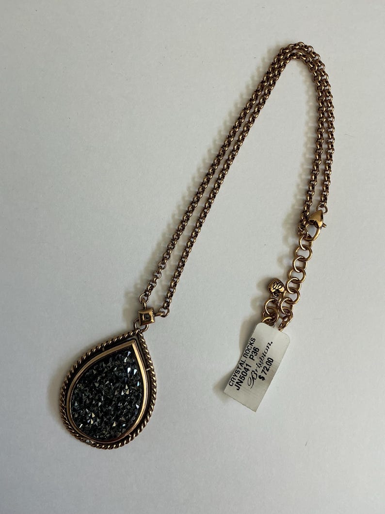 May include: A copper-toned necklace with a teardrop pendant filled with dark gray crystals. The pendant is framed with a gold-colored border and hangs from a matching chain. A tag with text is attached to the chain.