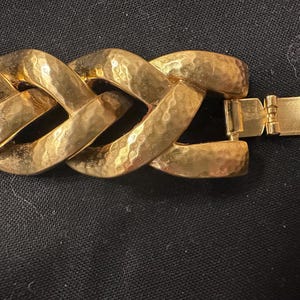 May include: Close-up of a gold-toned bracelet with a textured, hammered finish. The bracelet features large, interlocking links and a secure clasp. The jewelry is displayed against a dark background.