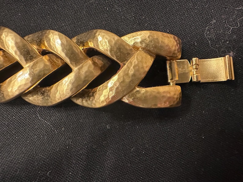 May include: Close-up of a gold-toned bracelet with a textured, hammered finish. The bracelet features large, interlocking links and a secure clasp. The jewelry is displayed against a dark background.