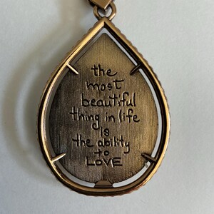 May include: A bronze-colored teardrop pendant necklace. The pendant has a textured interior with the engraved words "the most beautiful thing in life is the ability to LOVE". The pendant is framed with a beaded edge and hangs from a delicate chain.