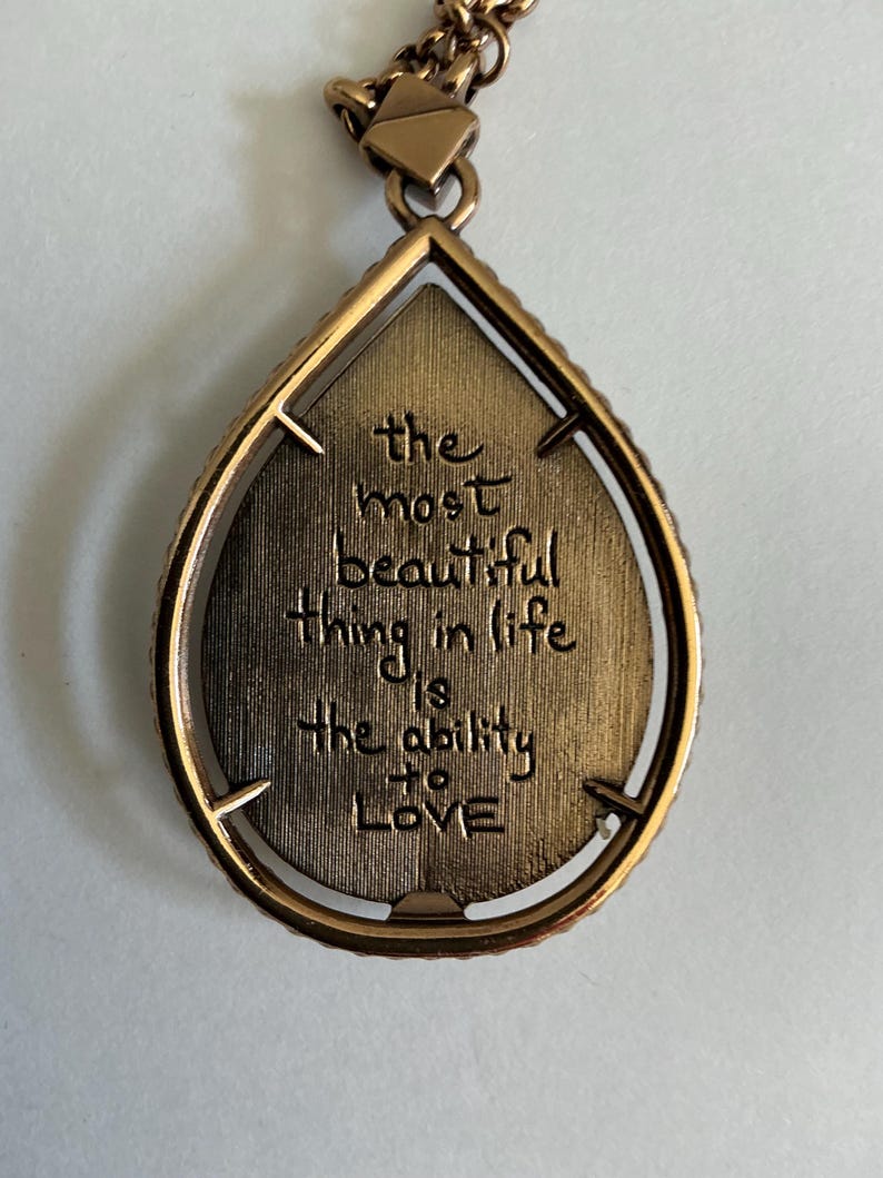 May include: A bronze-colored teardrop pendant necklace. The pendant has a textured interior with the engraved words "the most beautiful thing in life is the ability to LOVE". The pendant is framed with a beaded edge and hangs from a delicate chain.