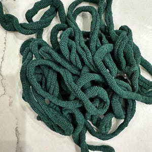 May include: A tangled pile of dark green rope, likely made of a knitted or woven material. The rope is thick and appears to be flexible, with a textured surface. The background is a white and gray marbled surface.