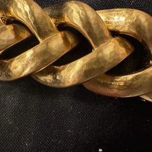 May include: Close-up of a gold-colored chain necklace. The chain links are large, rectangular, and have a hammered texture. The necklace is displayed against a black background, highlighting the gold color and the chain's design.