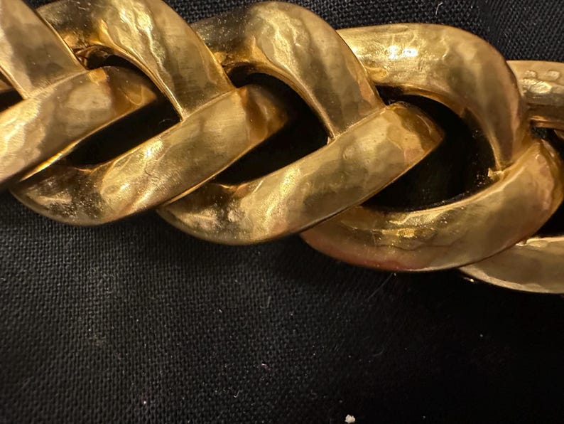 May include: Close-up of a gold-colored chain necklace. The chain links are large, rectangular, and have a hammered texture. The necklace is displayed against a black background, highlighting the gold color and the chain's design.