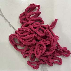 May include: A tangled pile of bright pink knitted fabric, possibly rope or cord, lies on a white surface with a gray vein pattern. The fabric is thick and appears to be made of a soft material.
