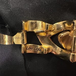May include: Close-up of a gold-toned metal clasp with the engraved text "Brighton" and "Live Life". The clasp is set against a dark background, highlighting its intricate design and polished finish. The clasp appears to be part of a bracelet or necklace.