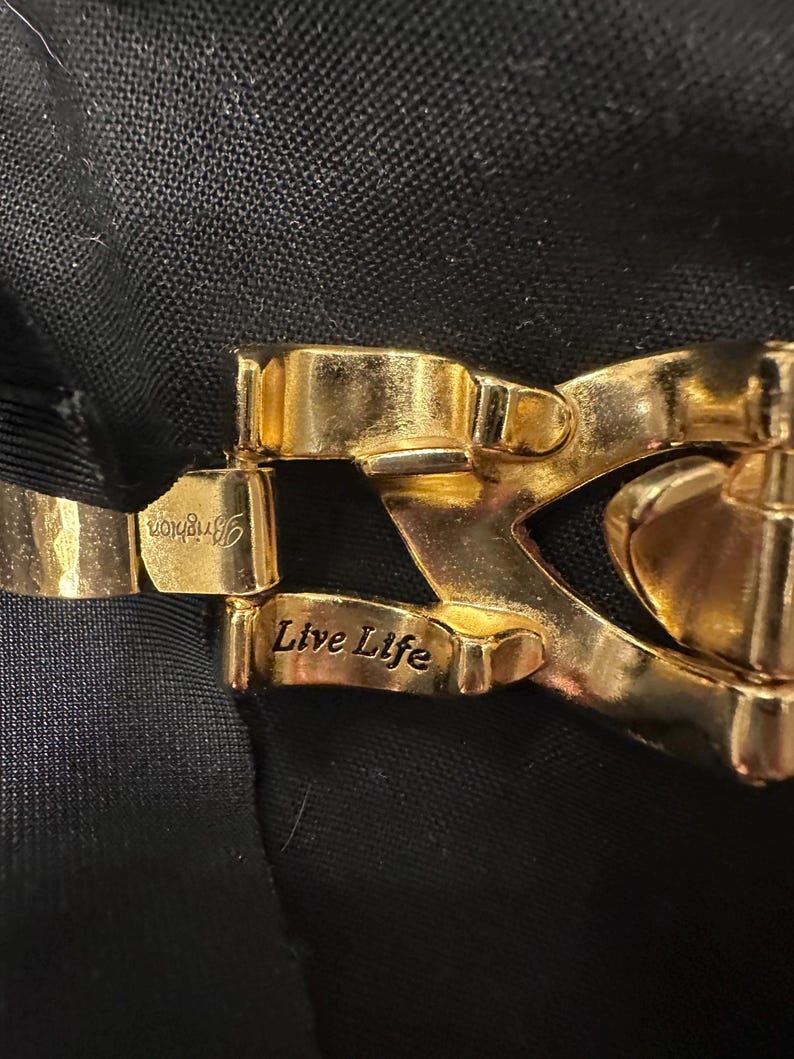 May include: Close-up of a gold-toned metal clasp with the engraved text "Brighton" and "Live Life". The clasp is set against a dark background, highlighting its intricate design and polished finish. The clasp appears to be part of a bracelet or necklace.