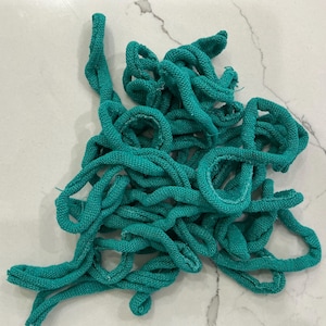 May include: A tangled pile of teal-colored, knitted rope. The rope is thick and appears to be made of a soft, textured material. The loops and curves of the rope create a visually interesting pattern against a white marble surface.