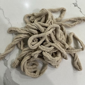 May include: A tangled pile of beige knitted rope lies on a light-colored surface. The rope appears to be made of a thick, textured yarn, with visible loops and knots. The rope is in a loose, unstructured arrangement.