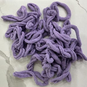 May include: A pile of lavender-colored knitted fabric tubes, arranged in a tangled heap. The tubes have a textured, open weave, and are of varying lengths and thicknesses. The fabric appears soft and pliable, suggesting a craft or textile project.