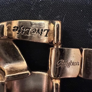 May include: Close-up of a gold-toned bracelet with the words "Live Life" and "Brighton" engraved on the links. The bracelet has a polished finish and is set against a dark background.
