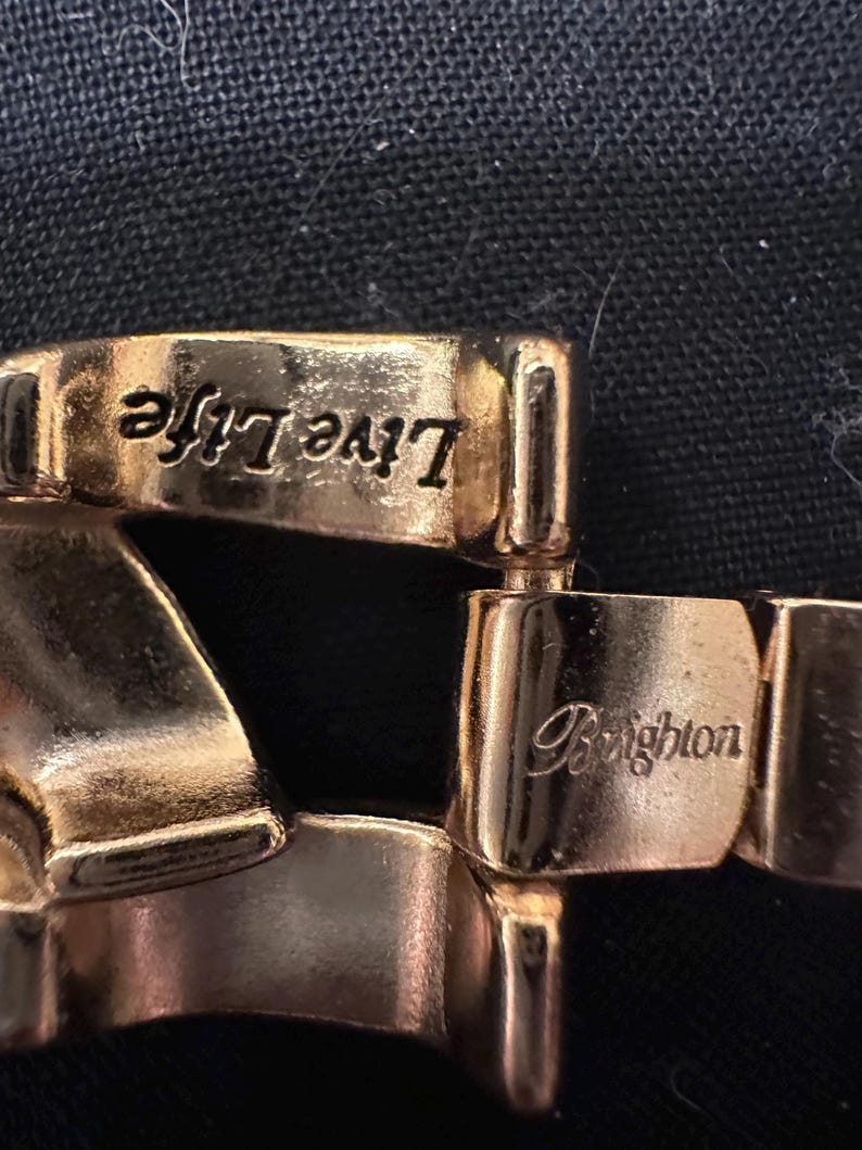 May include: Close-up of a gold-toned bracelet with the words "Live Life" and "Brighton" engraved on the links. The bracelet has a polished finish and is set against a dark background.