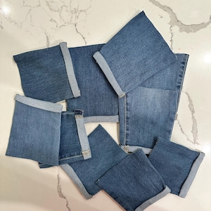 May include: A collection of denim squares, each with a folded cuff, arranged on a white marble surface. The squares are various sizes and shades of blue, showcasing the texture and color variations of the denim material. The arrangement suggests a craft or DIY project.