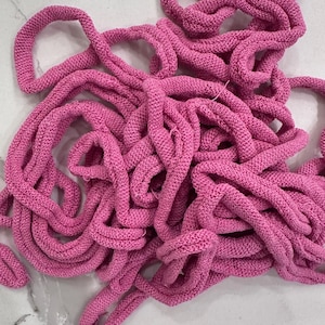 May include: A tangled pile of bright pink knitted cord. The cord is thick and appears to be made of a soft, textured material. The loops and curves of the cord create a visually interesting pattern against a white marble surface.