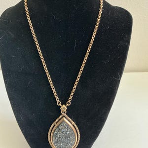 May include: A gold-toned necklace with a teardrop pendant. The pendant features a textured, gray stone framed by a twisted rope design. The chain is a delicate link style, and the necklace is displayed on a black mannequin.