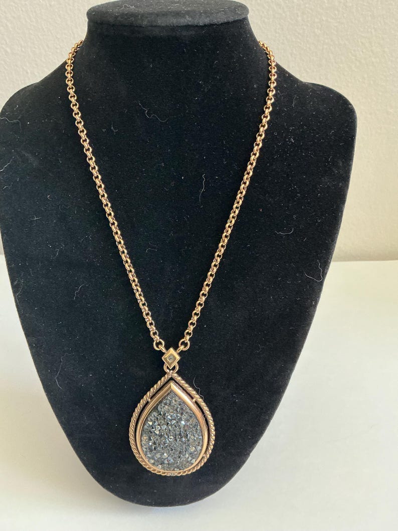May include: A gold-toned necklace with a teardrop pendant. The pendant features a textured, gray stone framed by a twisted rope design. The chain is a delicate link style, and the necklace is displayed on a black mannequin.
