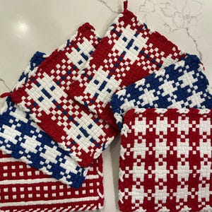 May include: A collection of woven potholders in red, white, and blue. The potholders feature geometric patterns and are arranged in a stack. Each potholder has a loop for hanging. The potholders are made of a thick, durable material.
