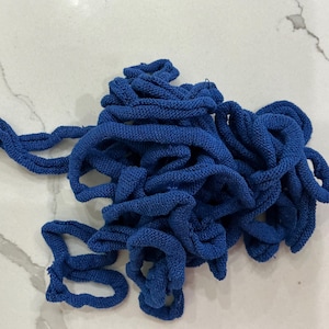 May include: A tangled pile of vibrant blue knitted fabric, possibly yarn or cord, lies on a white and gray marbled surface. The fabric appears soft and textured, with visible loops and a consistent color throughout.