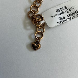 May include: Close-up of a gold-toned necklace with a heart-shaped charm. The necklace features a series of interlocking circular links. A tag is attached to the necklace. The heart charm has an engraved design.