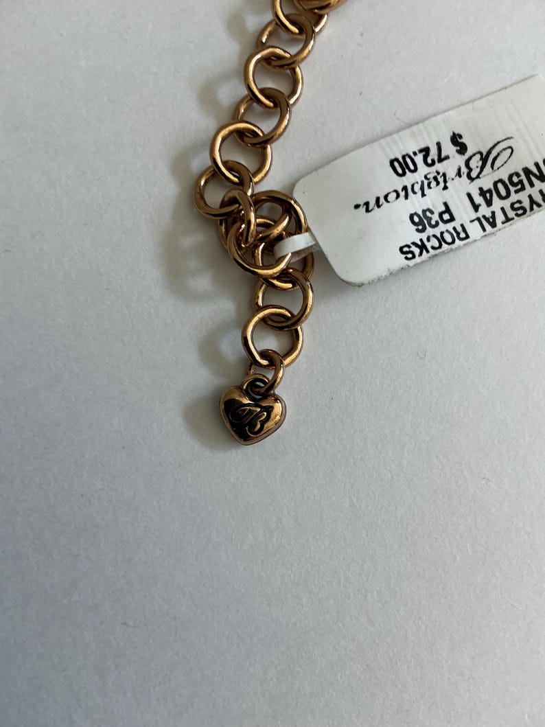 May include: Close-up of a gold-toned necklace with a heart-shaped charm. The necklace features a series of interlocking circular links. A tag is attached to the necklace. The heart charm has an engraved design.