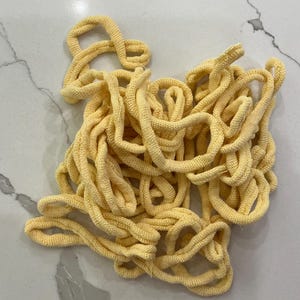 May include: A tangled pile of yellow knitted rope, possibly for crafting or decorative purposes. The rope is a light, pastel yellow color and appears to be made of a soft, textured material. The rope is lying on a white and gray marble surface.