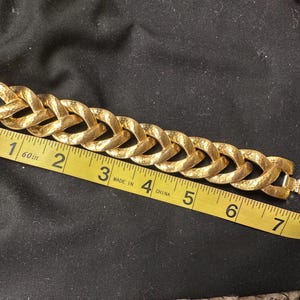 May include: A gold-toned bracelet with a textured, woven chain design. The bracelet is measured with a yellow measuring tape, showing a length of approximately 7 inches. The clasp is visible, and the bracelet is set against a dark background.