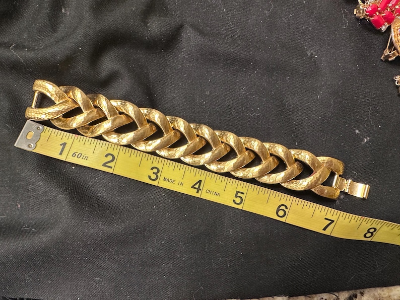 May include: A gold-toned bracelet with a textured, woven chain design. The bracelet is measured with a yellow measuring tape, showing a length of approximately 7 inches. The clasp is visible, and the bracelet is set against a dark background.