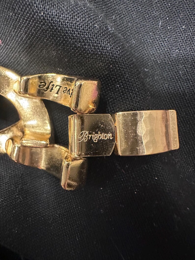 May include: Close-up of a gold-toned bracelet clasp. The clasp is rectangular with the brand name "Brighton" engraved on it. The bracelet links are also gold-toned, with the words "Live the Life" engraved on one link.