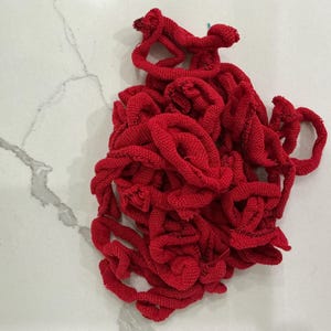 May include: A tangled pile of bright red knitted fabric, possibly yarn or cord, lies on a white and gray marbled surface. The fabric is tightly woven, with visible loops and textures. The overall shape is irregular and organic.