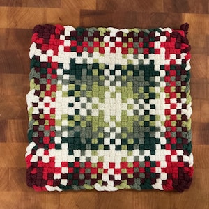 May include: A square, woven hot pad with a geometric pattern. The design features a central square of green and white, surrounded by a border of red, white, and green squares. The edges are finished with a burgundy trim.