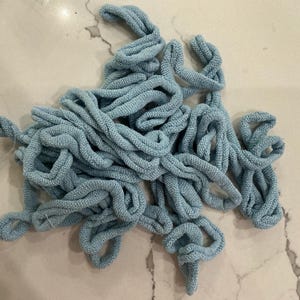 May include: A pile of light blue knitted fabric cords, tightly twisted and looped. The cords are a solid color and appear to be made of a soft, textured material. The cords are scattered across a light-colored surface.