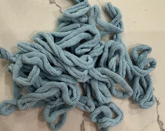 May include: A pile of light blue knitted fabric cords, tightly twisted and looped. The cords are a solid color and appear to be made of a soft, textured material. The cords are scattered across a light-colored surface.