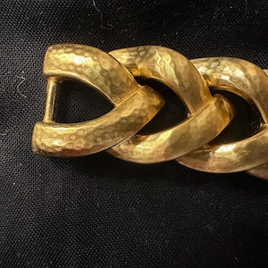May include: Close-up of a gold-toned chain link belt. The links are thick and textured with a hammered finish. The belt has a sturdy clasp. The background is a dark fabric.