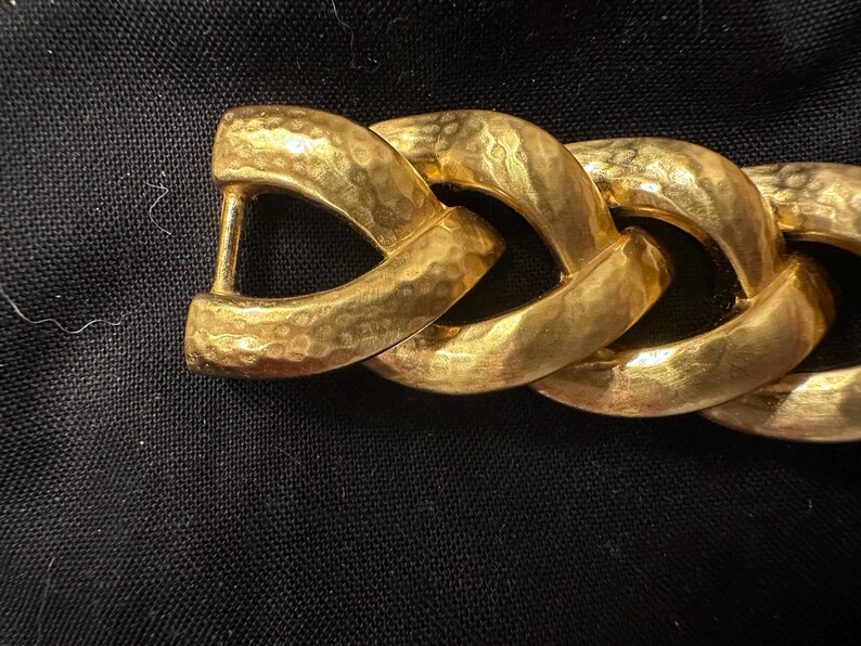 May include: Close-up of a gold-toned chain link belt. The links are thick and textured with a hammered finish. The belt has a sturdy clasp. The background is a dark fabric.