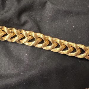 May include: A gold-colored bracelet with a textured, hammered finish. The bracelet features a series of interlocking oval links, creating a chain-like design. The clasp is a simple, rectangular design. The bracelet is a jewelry item.