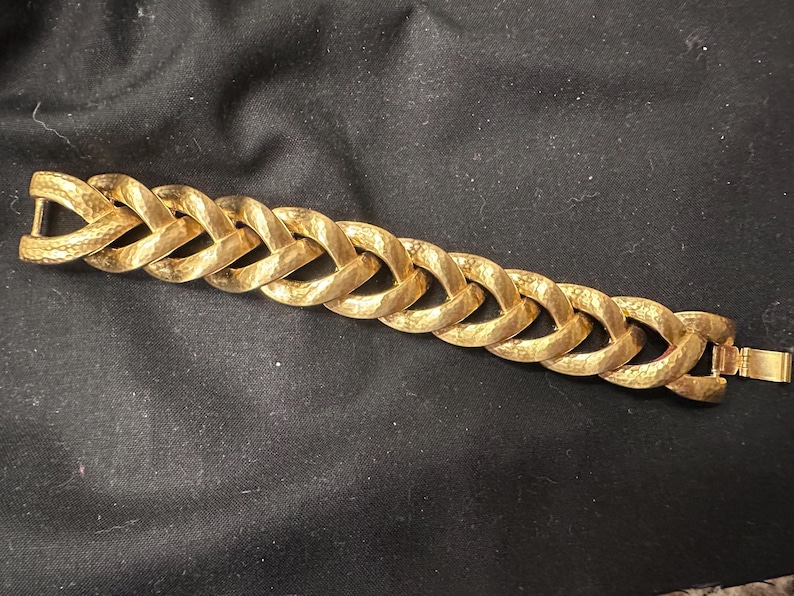 May include: A gold-colored bracelet with a textured, hammered finish. The bracelet features a series of interlocking oval links, creating a chain-like design. The clasp is a simple, rectangular design. The bracelet is a jewelry item.