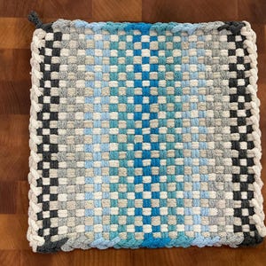 May include: Square woven potholder in shades of blue, grey, and black. The potholder has a braided edge and a loop for hanging. The woven pattern creates a textured surface, ideal for kitchen use.