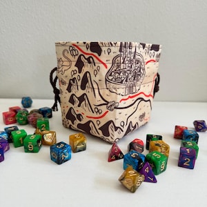 May include: A beige dice bag with a map design, featuring a castle and red lines, sits among scattered polyhedral dice. The bag has a drawstring closure. Dice are various colors, including green, blue, and gold.